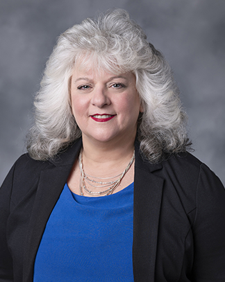 A professional headshot of Ann-Marie Corriveau, a smiling woman with shoulder-length gray hair, wearing a black blazer, a royal blue blouse, and sitting against a light gray background. The subject is facing the camera with a friendly and approachable expression.