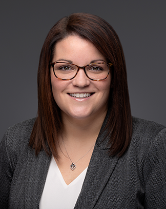 A professional headshot of Jill Sorel, a smiling woman with shoulder-length brown hair, wearing a gray blazer and sitting against a dark gray background. The subject is facing the camera with a friendly and approachable expression.